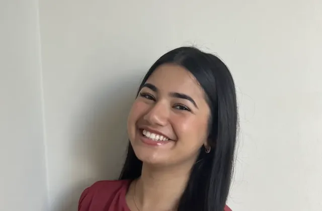 A woman with long dark hair wearing a sleeveless maroon top smiles while standing against a plain light-colored wall.