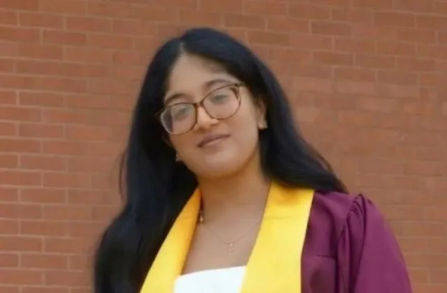 A young woman with long dark hair and glasses wearing a maroon graduation gown and yellow stole in front of a brick wall.