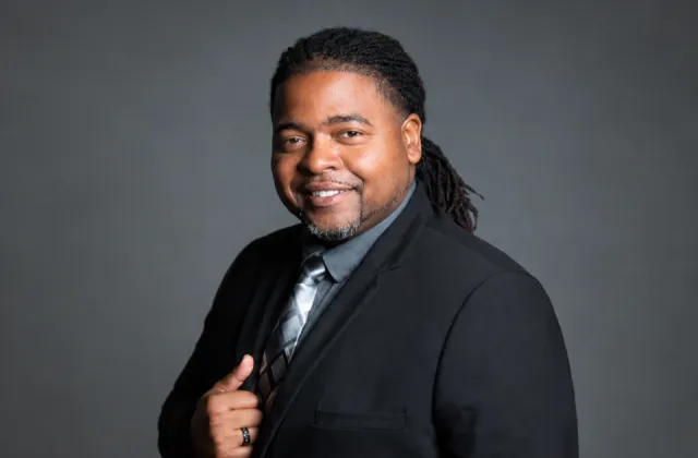 A man in a black suit, grey shirt, and patterned tie smiling at the camera.