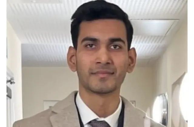 A professional headshot of a man in a beige blazer, white shirt, and tie.