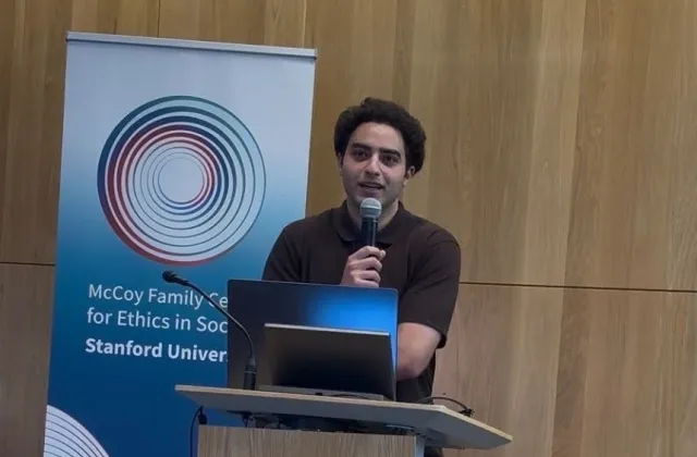 A man speaks into a microphone at a podium in front of a Stanford McCoy Family Center for Ethics in Society banner.