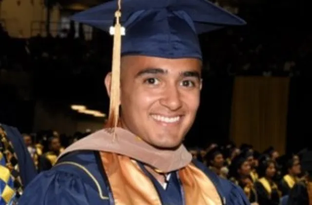 A smiling graduate in a blue cap and gown with a gold stole at a commencement ceremony.