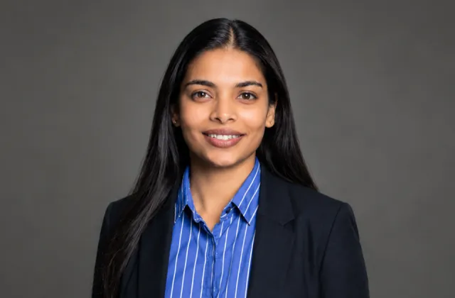 Professional portrait of a woman with long dark hair wearing a blue striped shirt and a black blazer against a grey background.