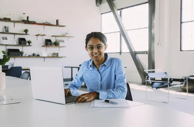 A woman wearing glasses sits at a desk with a laptop and documents in a bright, modern office space.