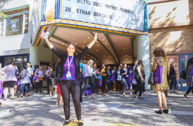 A woman stands smiling with arms raised in front of a theater entrance, surrounded by a crowd of people, under a marquee sign displaying event information.