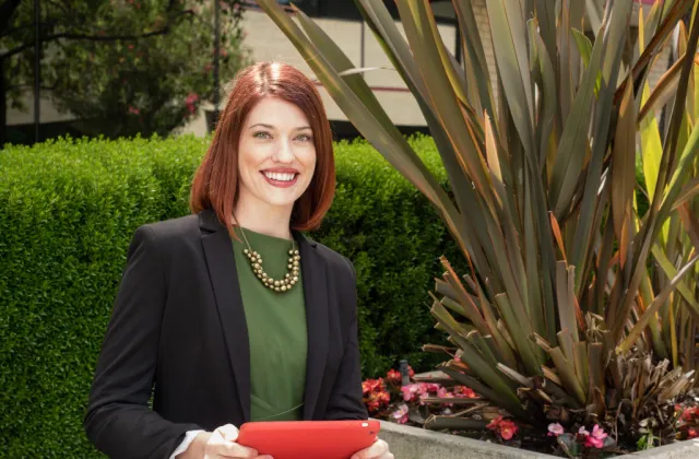 A woman with red hair, wearing a black blazer and green blouse, sits at an outdoor table holding a red tablet, with greenery and plants in the background.