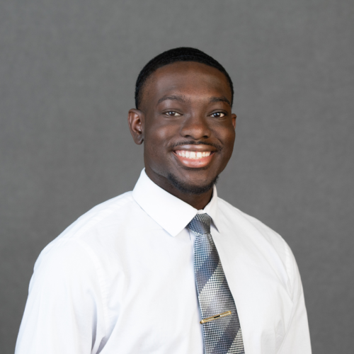 A man wearing a white dress shirt and patterned tie smiles at the camera against a plain gray background.