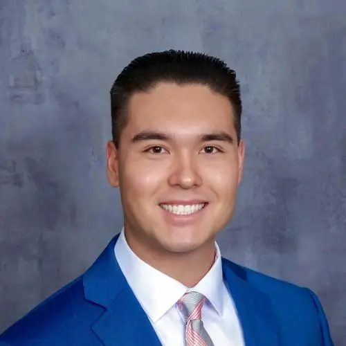 A young man wearing a blue suit, white shirt, and striped tie smiles in front of a gray mottled background.