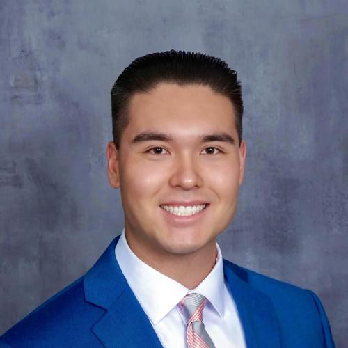 A young man wearing a blue suit, white shirt, and striped tie smiles in front of a gray mottled background.