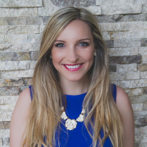 A woman with long blonde hair, wearing a blue sleeveless top and a floral necklace, smiles in front of a textured stone wall background.