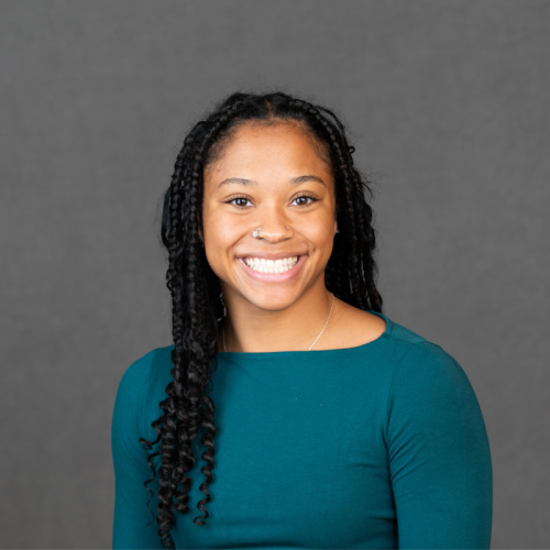 A woman with long braided hair, wearing a teal top, smiles at the camera against a plain gray background.