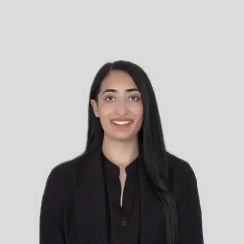 A woman with long dark hair, wearing a black blazer and dark shirt, stands in front of a plain light gray background, facing the camera and smiling.