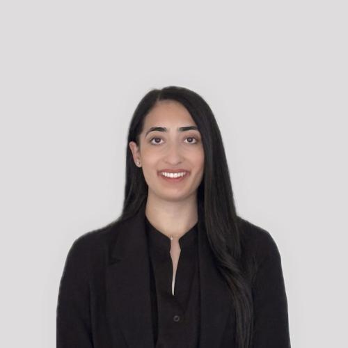 A woman with long dark hair, wearing a black blazer and dark shirt, stands in front of a plain light gray background, facing the camera and smiling.