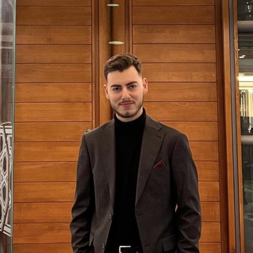A man in a dark suit and black turtleneck stands indoors in front of wooden panel doors, with his hands in his pockets, facing the camera.