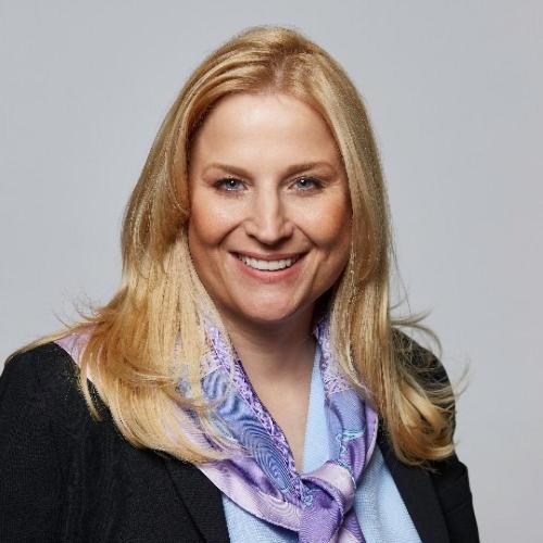 A woman with long blonde hair smiles, wearing a black blazer and a light purple patterned scarf, against a plain gray background.