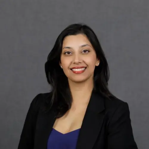 A woman with long dark hair wearing a black blazer and blue top smiles in front of a plain gray background.