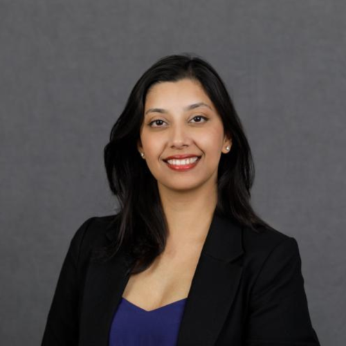 A woman with long dark hair wearing a black blazer and blue top smiles in front of a plain gray background.