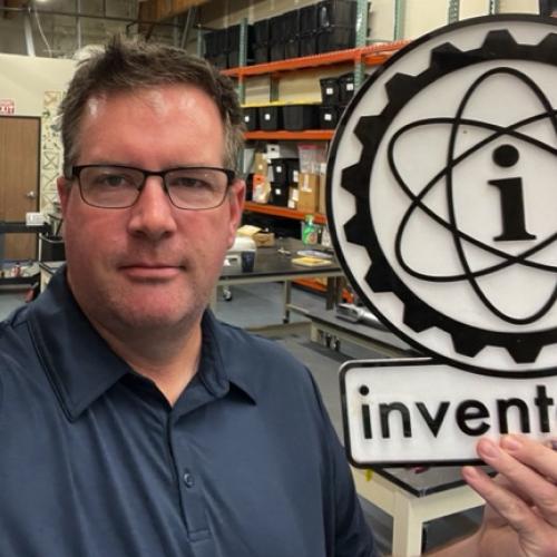 A man in a workshop holds a large sign with an atomic symbol and the word “inventopia.” Shelves and tools are visible in the background.