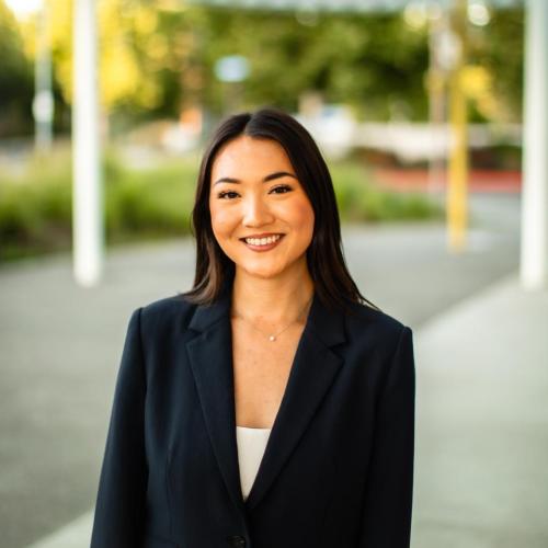A woman in a dark blazer stands outdoors, smiling at the camera, with a blurred background of greenery and architectural structures.
