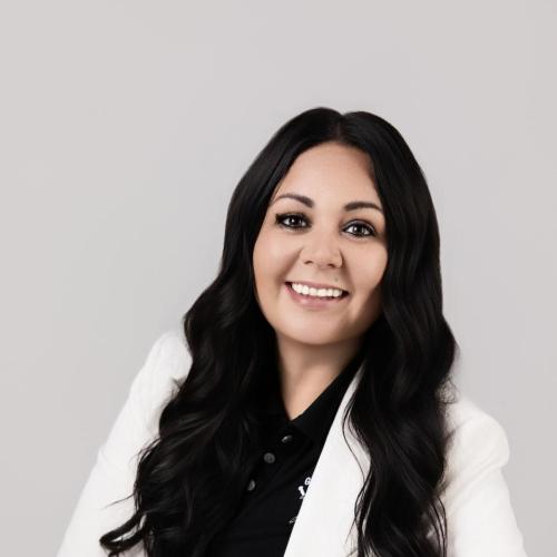 A woman with long dark hair, wearing a white blazer over a black top, is seated and smiling at the camera against a plain light background.