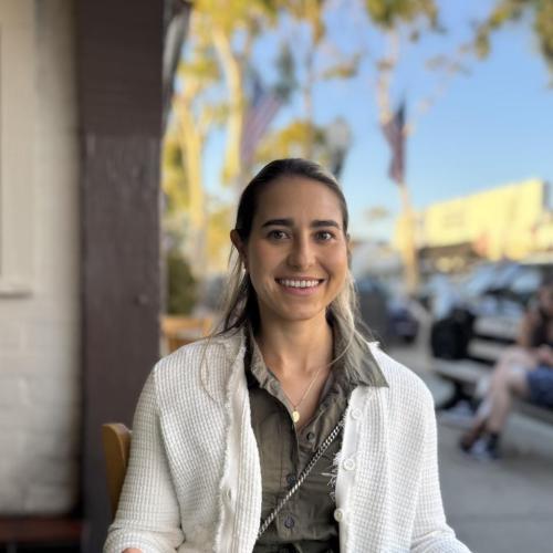 A woman with long hair, wearing a white sweater over a green shirt, sits outdoors at a restaurant table, smiling at the camera. Trees and parked cars are visible in the background.