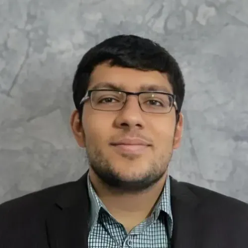 A man wearing glasses and a checked shirt with a dark blazer is posed in front of a gray textured background.