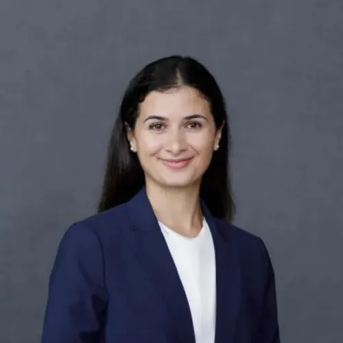 Woman with long dark hair wearing a navy blazer and white blouse, smiling in front of a plain gray background.