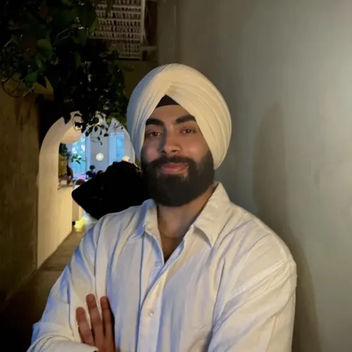 A person wearing a white turban and white shirt stands indoors with arms crossed, facing the camera and smiling slightly.