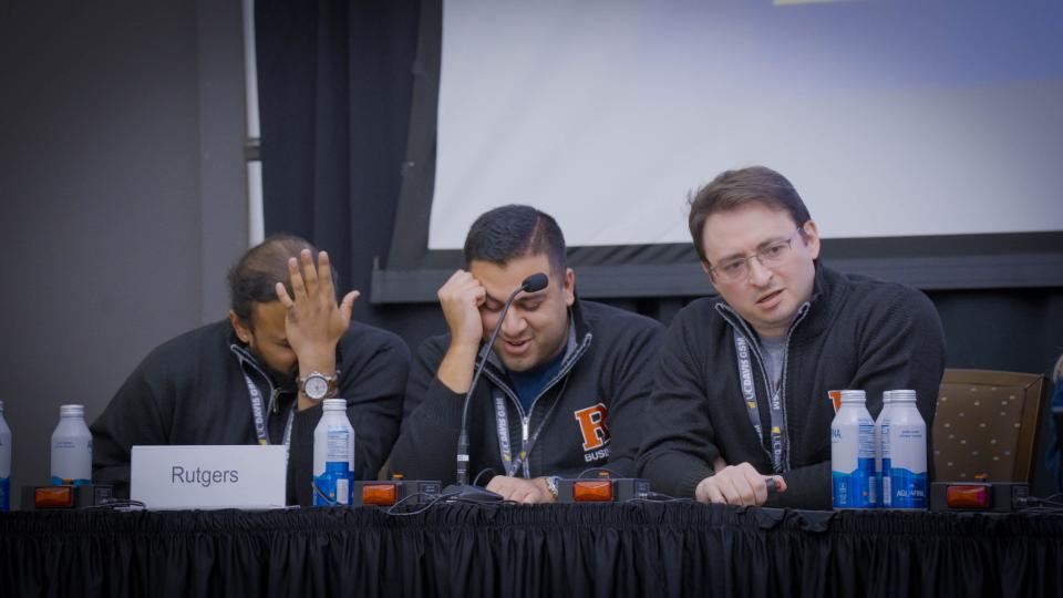 Three men sitting at a table with a mic and buzzers placed in front of them