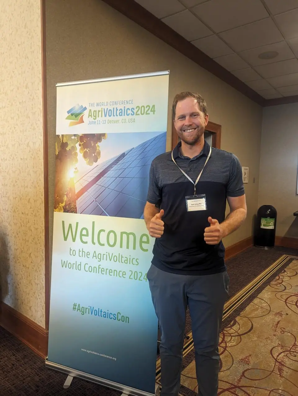 Jack Schaufler holding up two thumbs up, standing next to a welcome sign at a conference