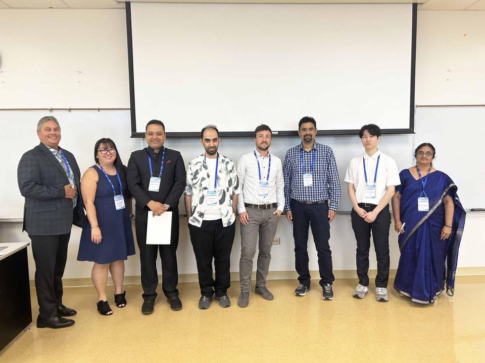 A group of eight international conference presenters and organizers standing in a classroom in front of a whiteboard and projector screen. They are wearing conference badges and formal or business casual attire.