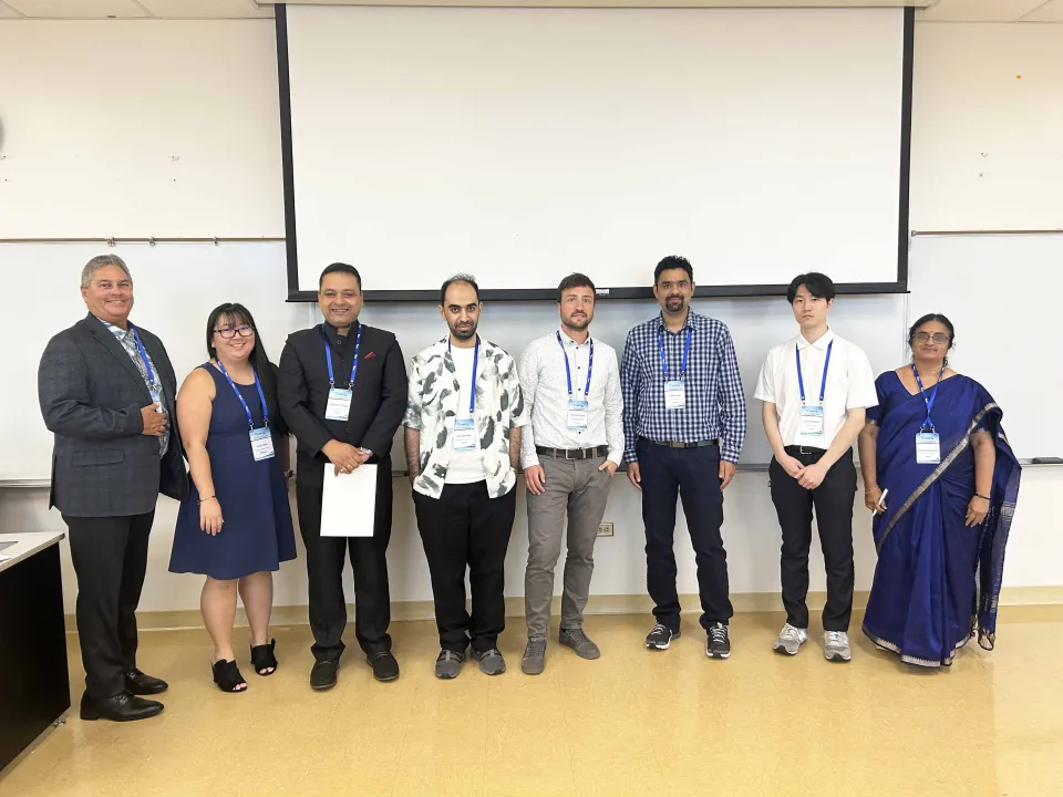 A group of eight international conference presenters and organizers standing in a classroom in front of a whiteboard and projector screen. They are wearing conference badges and formal or business casual attire.