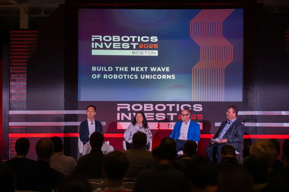 Koji Hasegaw with panelists speak onstage at Robotics Invest 2025 in Boston, seated in front of a large screen that reads “Build the Next Wave of Robotics Unicorns.” Audience members watch from the foreground.