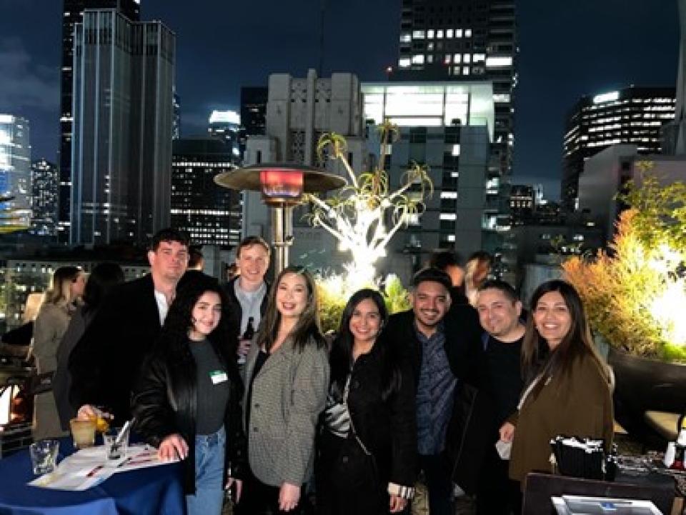 A group of UC Davis Graduate School of Management students and alumni gather on a rooftop patio at night, smiling together with the city skyline lit up behind them.