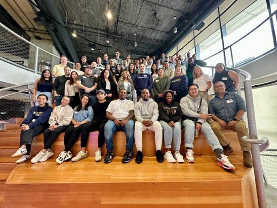 A large group of UC Davis MBA students and faculty pose together on wooden stadium-style steps inside a modern building, smiling and gathered closely as part of a weekend residential event.