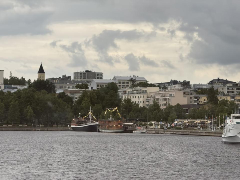 Harborfront in Lappeenranta, Finland, with boats docked along the shoreline and apartment buildings rising behind under a cloudy summer sky.