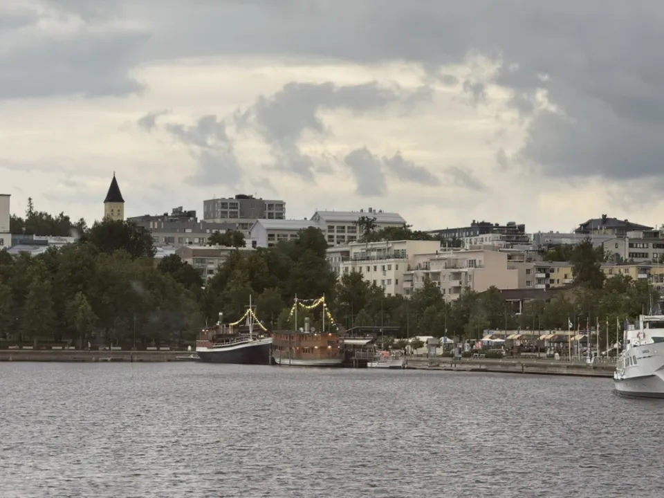 Harborfront in Lappeenranta, Finland, with boats docked along the shoreline and apartment buildings rising behind under a cloudy summer sky.