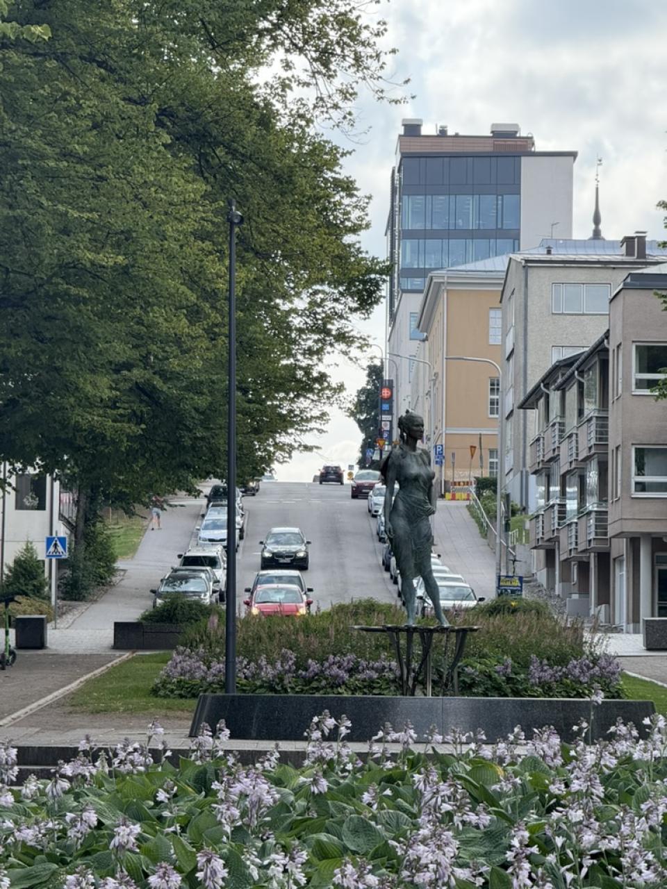 Street view in Lappeenranta, Finland, featuring a bronze statue in the foreground, flowerbeds, and a tree-lined road leading uphill past modern and historic buildings.