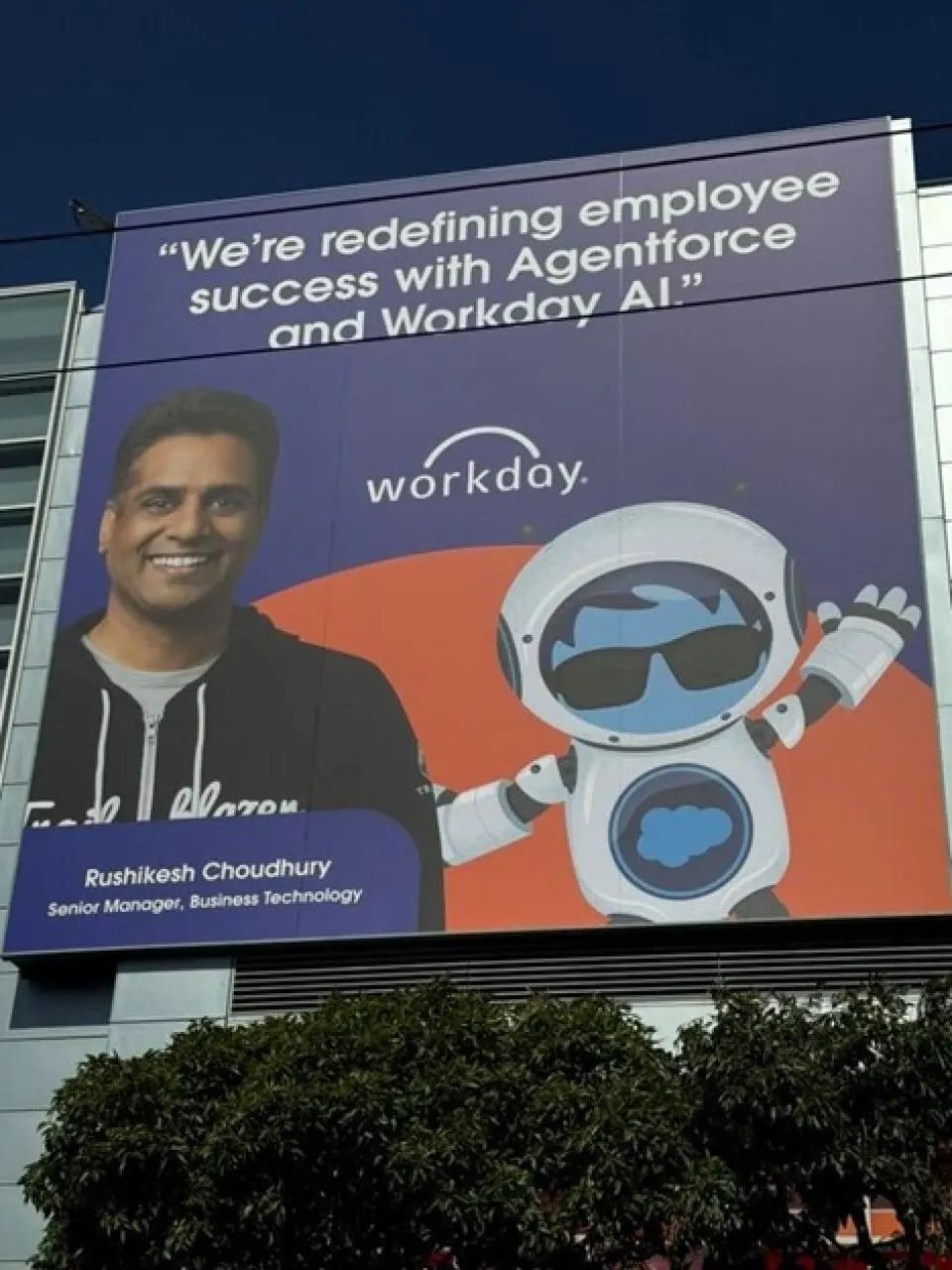 Large billboard featuring Rushikesh Choudhury, Senior Manager of Business Technology at Workday, alongside a cartoon robot. The text reads: “We’re redefining employee success with Agentforce and Workday AI.”