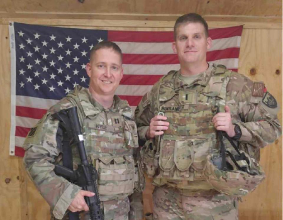 Nathaniel Wentland next to a soldier, both dressed in fatigues and standing in front of the American flag