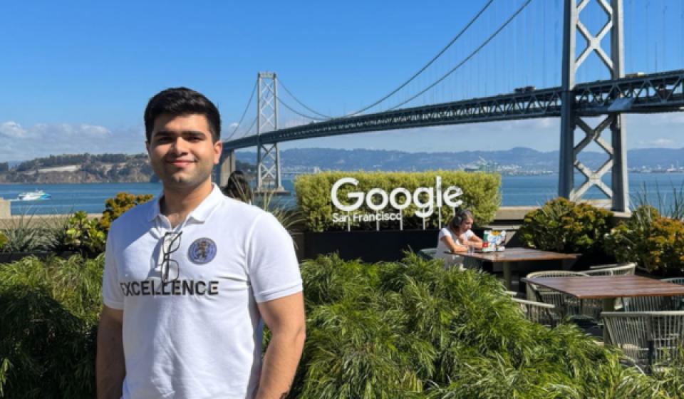 Talha Amin stands on an outdoor terrace at Google’s San Francisco office, with the Bay Bridge and blue sky in the background. He’s wearing a white polo shirt, light pants and sunglasses, surrounded by lush green plants.