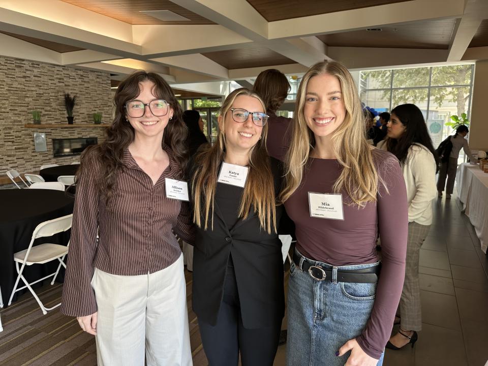 Three UC Davis MPAc students, including Mia Hildebrand on the right, stand together and smile at a networking event indoors.