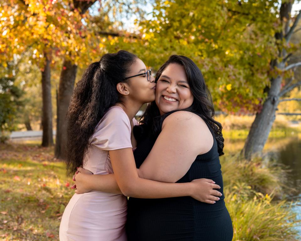 Melissa and her teenage daughter embrace outdoors in front of trees with fall leaves, one kissing the other on the cheek as they both smile.