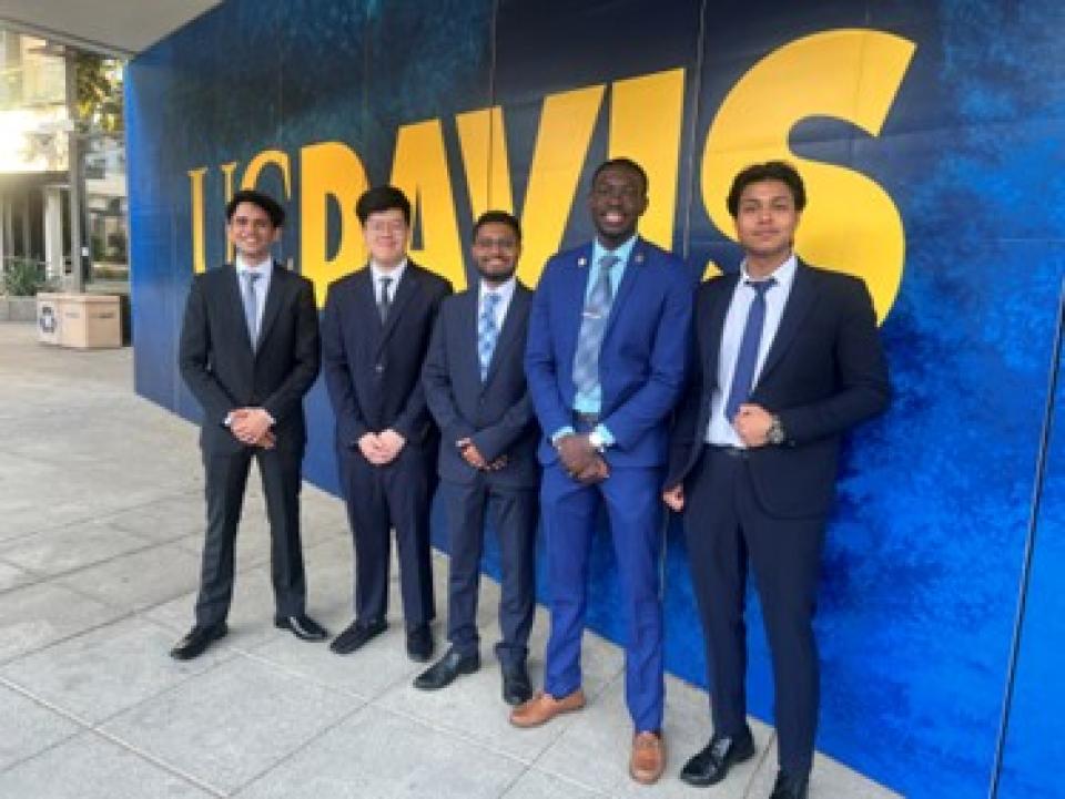 Five men in suits stand in front of a large UC Davis sign on a sidewalk, posing for a group photo.