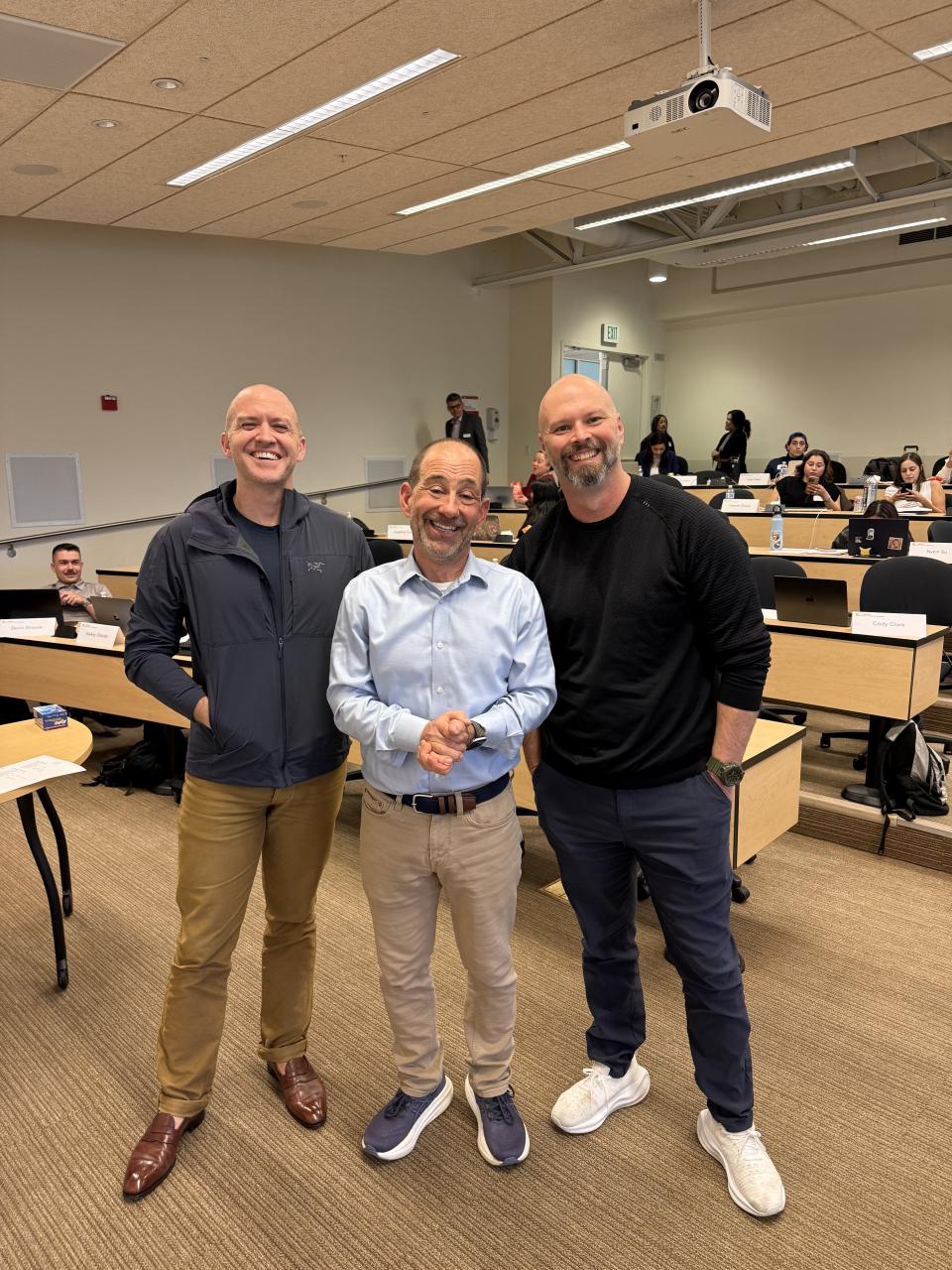 Three men standing and smiling in a classroom setting, with students seated at desks and a projector on the ceiling above them.