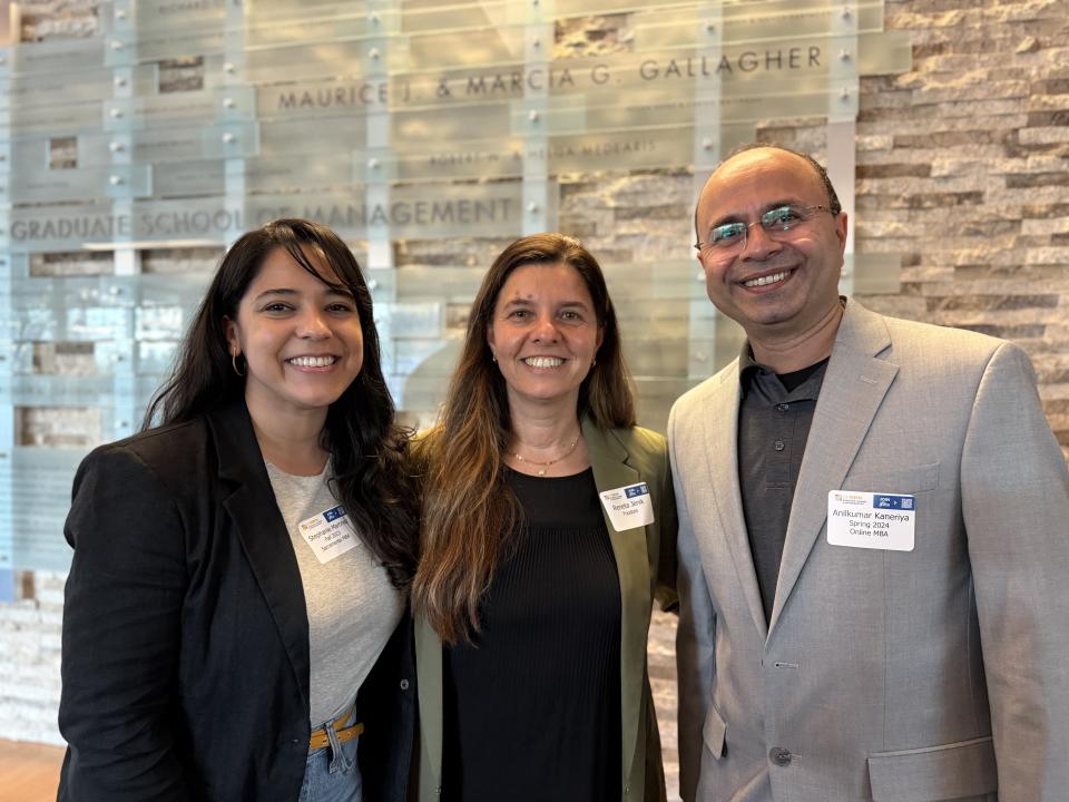 Three people stand indoors, smiling at the camera. They wear business attire and conference name tags. A wall sign behind them reads "Graduate School of Management.