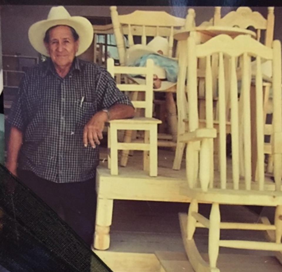 An older man in a cowboy hat stands next to handmade wooden chairs and rockers, displaying his craftsmanship.