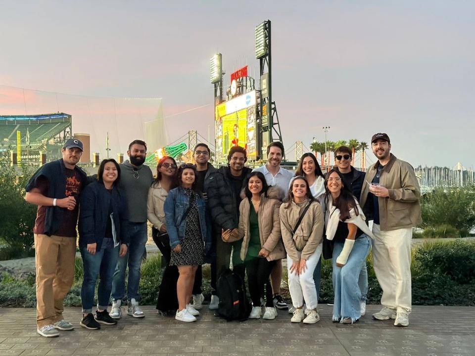 A group of fourteen people stand together outdoors in front of a baseball stadium scoreboard at dusk, smiling at the camera.
