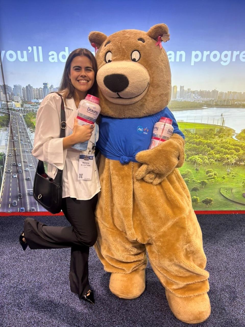 A woman smiles and poses with a person in a bear costume, both holding bottles of Downy Unstopables, in front of a scenic city backdrop.