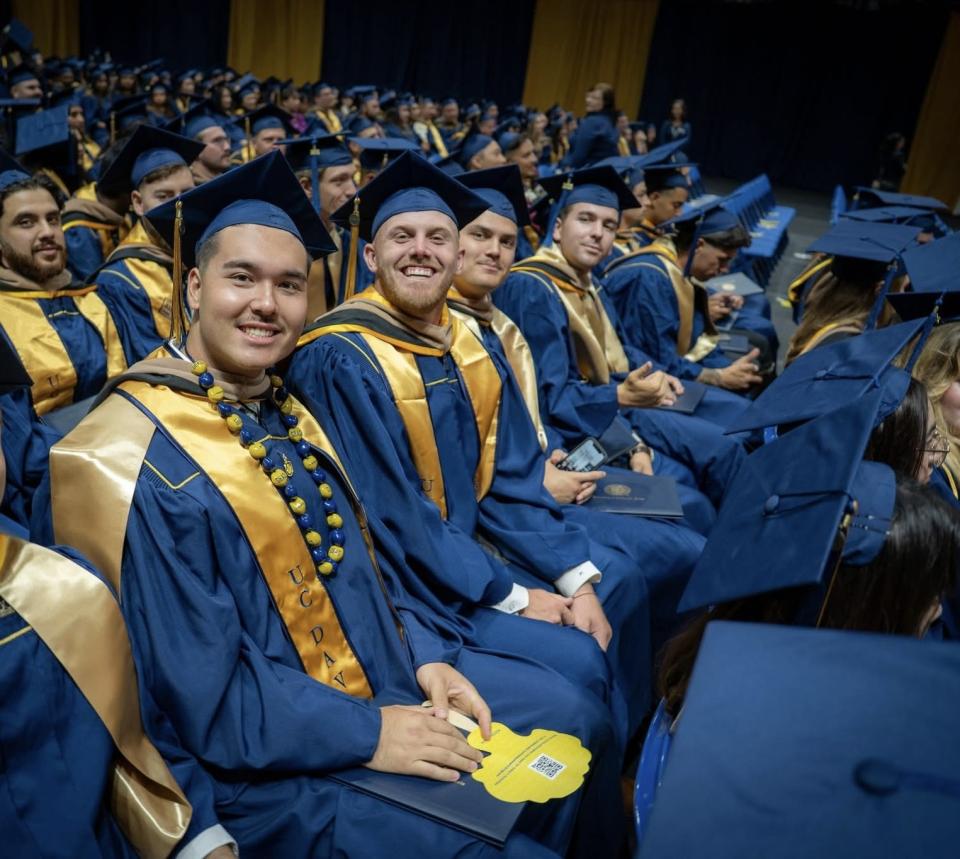 Graduates in blue caps and gowns with gold stoles sit in rows at a commencement ceremony, holding programs and smiling at the camera.